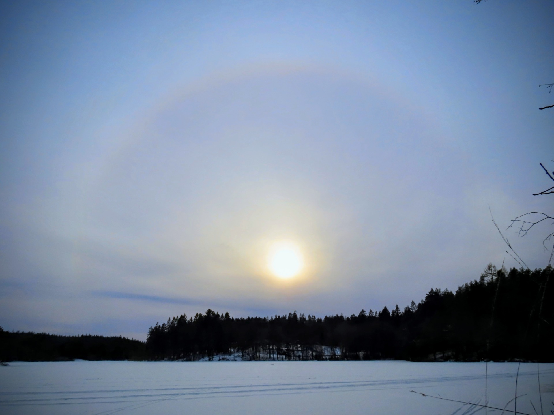Auringonlasku talvisen järvimaiseman yllä, metsän siluetti ja taivaalla pilviä.